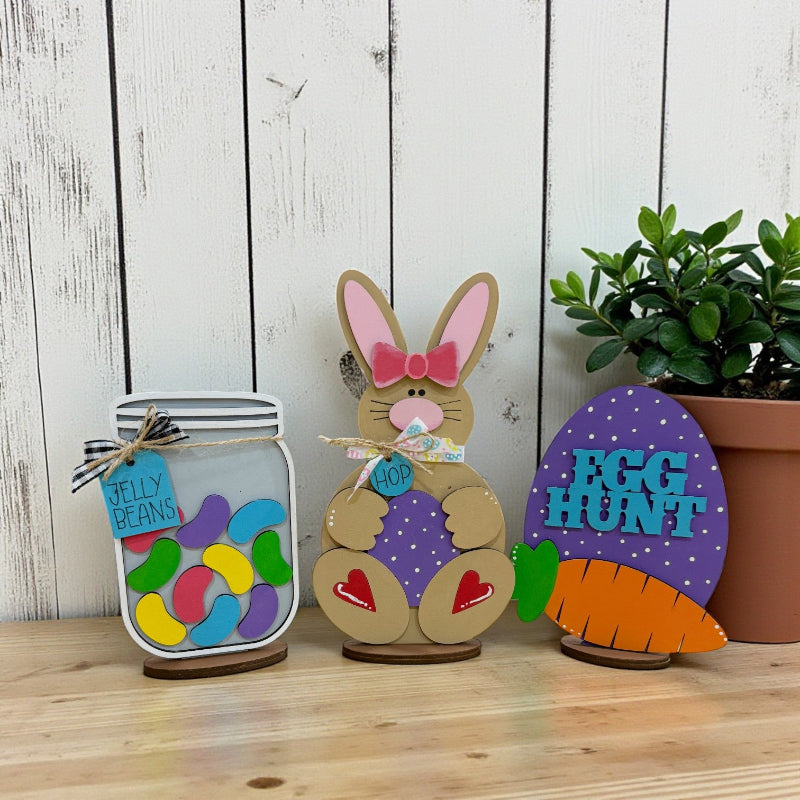 Hand-painted Easter mini stand inserts featuring jelly beans jar, bunny with egg, and Egg Hunt carrot layered wood designs displayed on tabletop by Jazzi’s Creations in Orange Park Florida