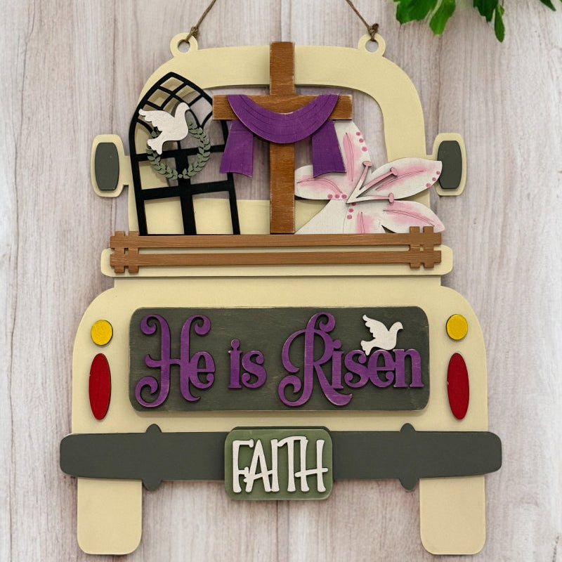 A wooden DIY truck insert with a 'He is Risen' sign, decorated with a cross, Easter lilies, a window with a dove, and a 'Faith' license plate, in a beige, purple, and green color scheme, designed for interchangeable Farmhouse Truck decor.