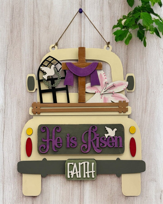A wooden DIY truck insert with a 'He is Risen' sign, decorated with a cross, Easter lilies, a window with a dove, and a 'Faith' license plate, in a beige, purple, and green color scheme, designed for interchangeable Farmhouse Truck decor.