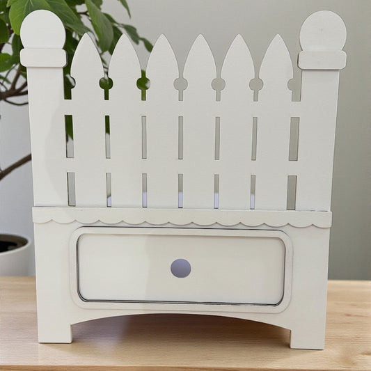 White picket fence-shaped planter on a wooden surface with a plant in the background.
