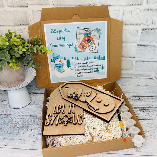 Wooden snowman tags and paint supplies in an open cardboard box on a wooden surface craft kit at jazzis creations in orange park fl