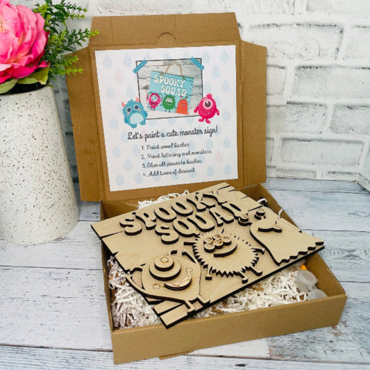 A Spooky Squad Halloween DIY Craft Kit for Kids is displayed, including a set of pre-cut wooden monster shapes, paint set, brushes, and a mess-free apron, arranged inside an open cardboard box at jazzis creations in orange park fl